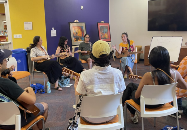 A group of people sits in a circle indoors, playing stringed instruments and listening to an instructor near a whiteboard. Art and a TV are visible on the walls.