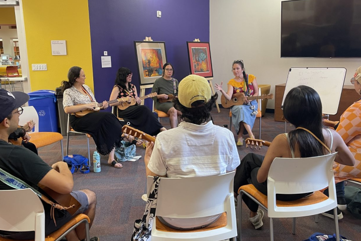 A group of people sits in a circle indoors, playing stringed instruments and listening to an instructor near a whiteboard. Art and a TV are visible on the walls.